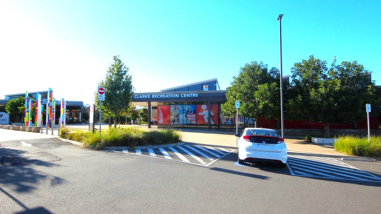 A white car is parked in one of the accessible parking lots in Michael Clarke Recreation Centre, marked by white lines and a blue accessible parking sign. A no-entry parking sign can be seen on the left side of the accessible parking area within the entry garden. Long but thin rectangular blocks with colorful designs are placed behind the sandstone blocks.