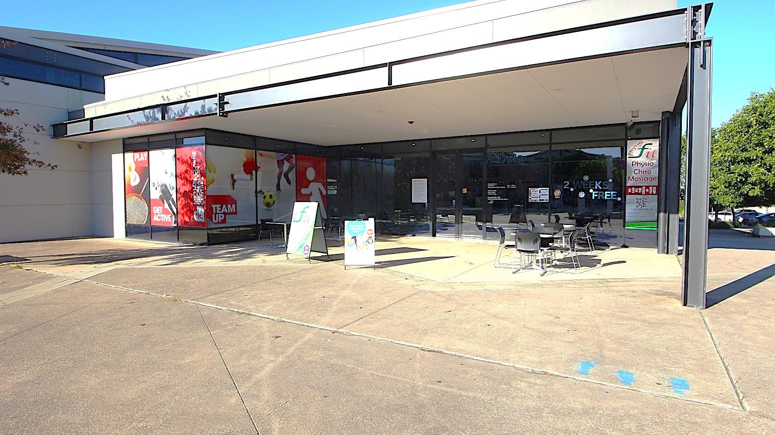 Entry to Michael Clarke Recreation Centre is by the tinted glass doors with black edges. The entrance can be distinguished by black metal frames, a seating area with white tables, and black with silver leg frames, chairs, and marking posters displayed on the tinted glass windows. Further to the back of the building are cars parked in the parking area.
