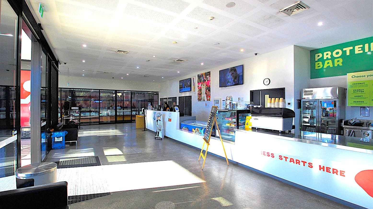 Overview of the entire reception area and Café as well the glass windows further to the left of the reception. The basketball center is visible through these glass windows. From this image, the double doors at the Michael Clarke Recreation Centre are automatic. There is a silver rubbish bin near the door, and several cold drinks stored in a large fridge which are available in the Café.