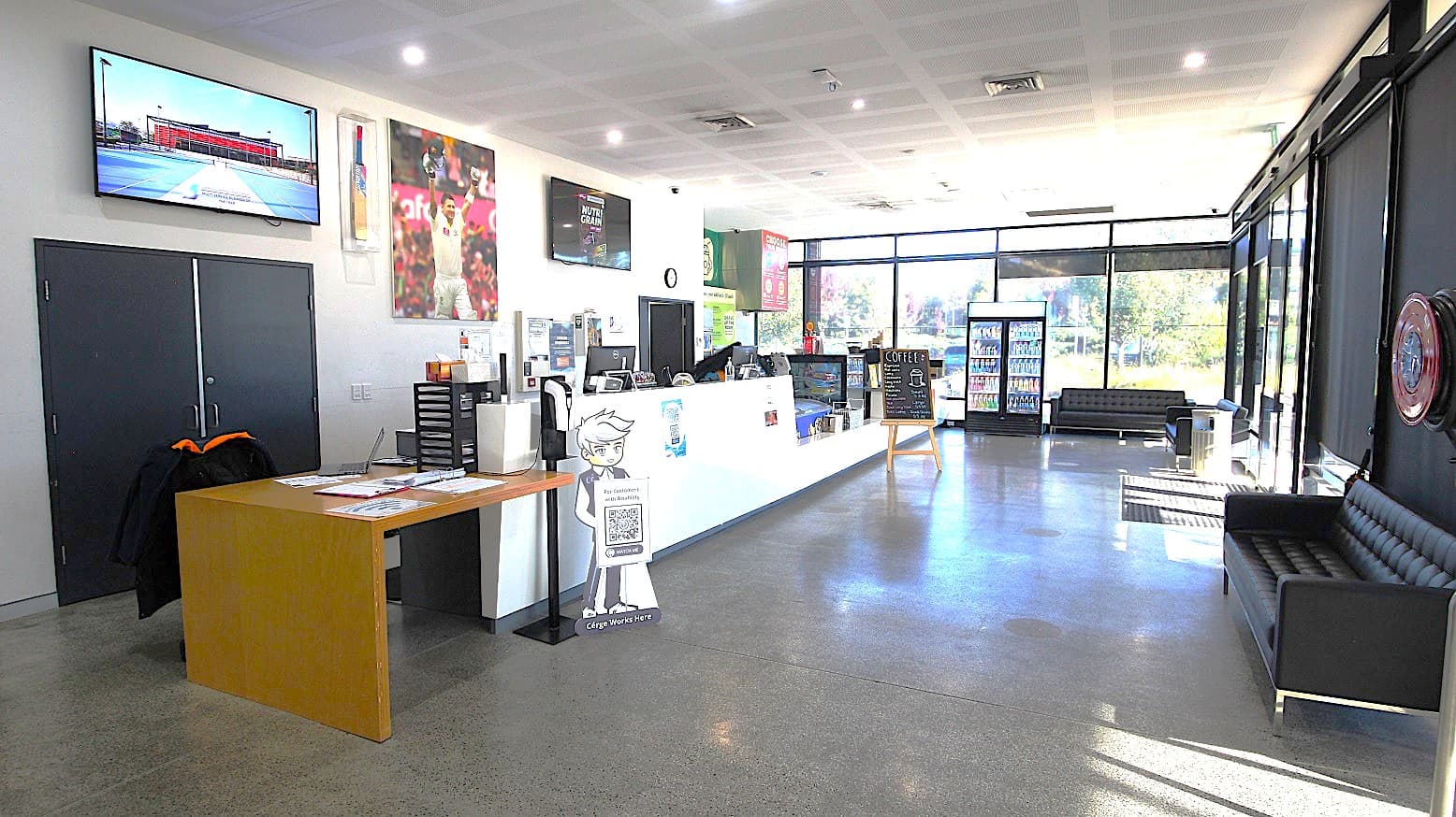 Opposite view of the entire reception area and Café from the previous image and the glass windows with the view of the natural scene on the outside. A television is mounted on the white wall at the reception desk, and underneath is a black door with metal handles. There is a brown wooden table at the end of the long reception counter to the left and an automatic alcohol dispenser beside the Cérge brand icon. Several sofa chairs are placed against the glass walls in this floor area.