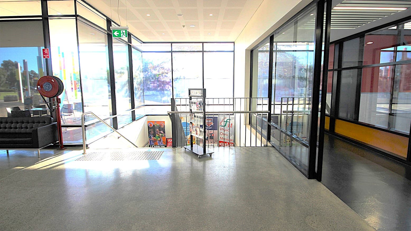 Black stairwell access to the gym and multipurpose rooms at the Michael Clarke Recreation Centre. Before the stairwell is a cart containing flyers that are arranged on different levels. Left of the staircase is a red fire hose mounted on the metal frame of the glass walls. On the right is a corridor space with black floors. Different marketing posters promoting the available amenities inside the venue are displayed further down the stairwell.