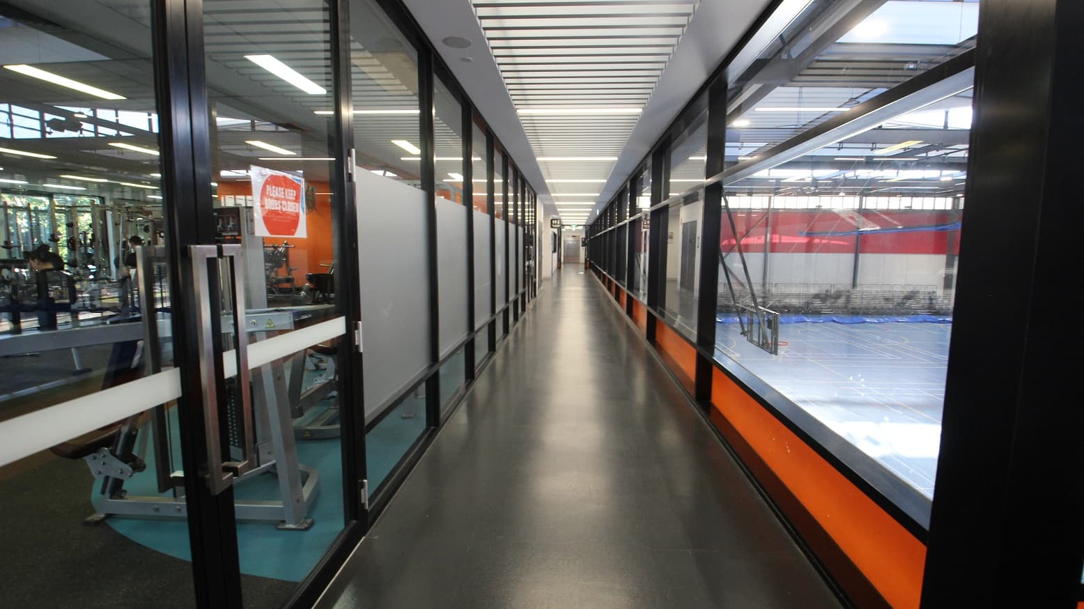 The entrance to the Gymnasium & Exercise Studio at Michael Clarke Recreation Centre is through a long corridor with glass walls and an orange silhouette at the lower portion, overlooking the Stadium. Entrance to the gym through a double door with grey metal handles requires a swipe card entry. The ceiling has a unique ladder-looking design.