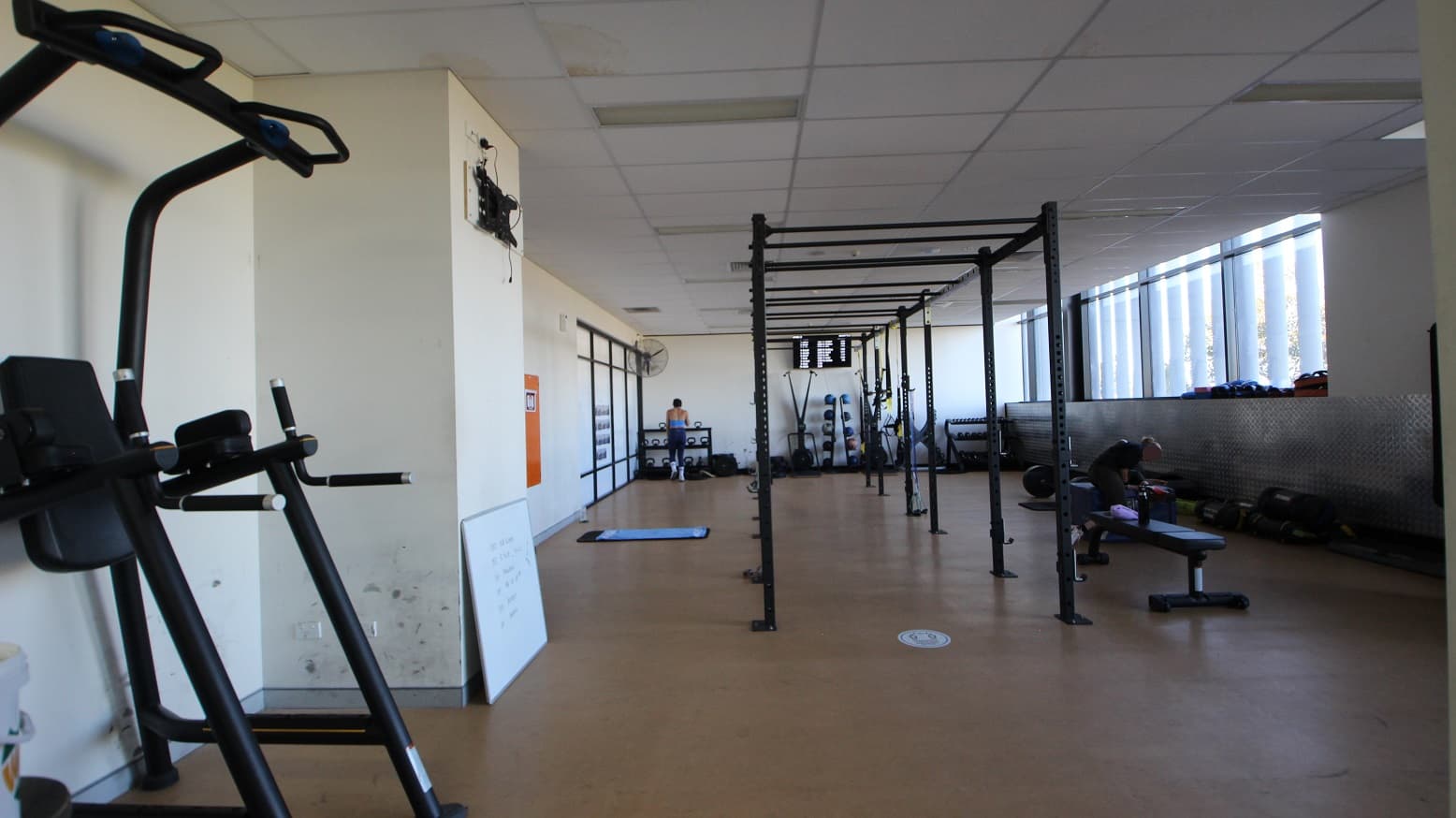 The image shows another part of the Gymnasium. The walls are painted white, and a shiny silver metal platform with a mid-waist height is placed against the right wall of the room. In the middle is a black crossfit rig and a bench. A medium size whiteboard is laid against the left wall, and different resistance training equipment is accessible further at the back.