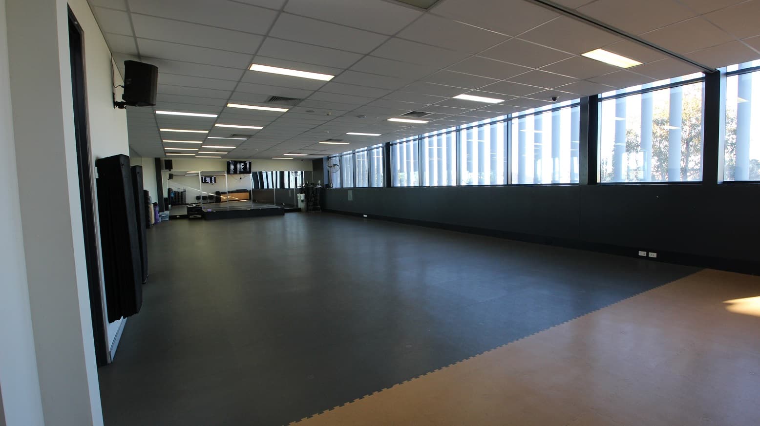 A room with a large open space covered with grey rubber mats in the Gymnasium of Michael Clarke Recreation Centre. Further to the back are floor-to-ceiling mirrors and a black elevated platform that looks like a small stage. On the left are speakers mounted on the wall.
