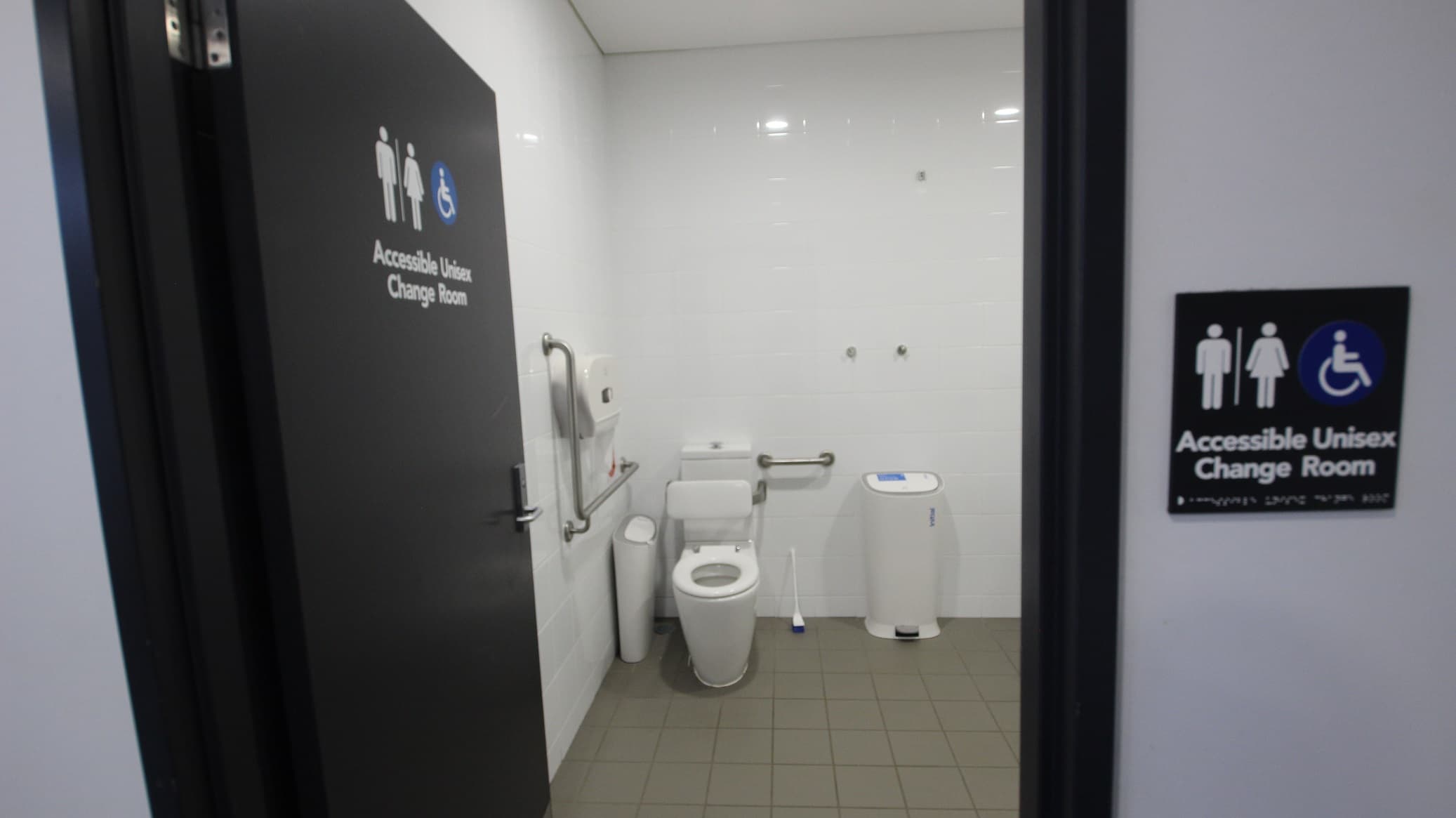 The image shows a part of the Accessible Unisex Change Room for persons with mobility disabilities in the Gymnasium. This change room can be distinguished by the blue accessible sign as well as male and female icons. The floor tiles are grey, while the wall tiles are white which makes the room polished and immaculate to the feeling. This image only shows the toilet bowl, white rubbish bin, toilet tissue paper holder, and blue toilet brush with white holder.