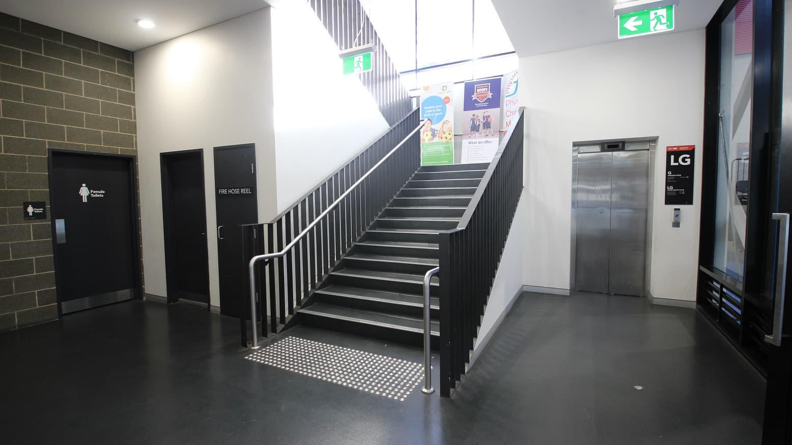 End of the black stairwell from the reception area. The stairwell has black metal handrails with a horizontal metal tube intersecting its parallel frame. On the left side are the black door of the Female Toilet within the grey brick wall and two small black doors with Fire Hose Reel labels within the white walls. On the right side is the elevator.