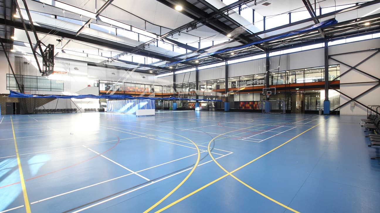 View from one of the corners of the Stadium at the Michael Clarke Recreation Centre. The stadium has blue floors with white, yellow, black, and red lines. The whole space in the Stadium is separated by black nets with blue cloth at the bottom. The natural lighting from the skylights is also supported by the artificial lighting coming from fluorescent bulbs. On the sides of the main floor of the Stadium are grey chairs.
