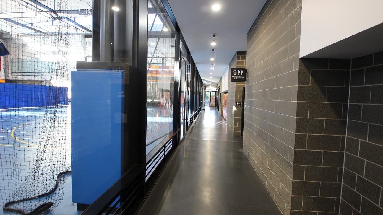 Corridor leading to Change Rooms and Showers at the Michael Clarke Recreation Centre. This corridor is separated from the Stadium by the glass walls and can be distinguished by its narrow area and grey brick walls. A number of CCTV cameras are also seen in the middle of the ceiling.