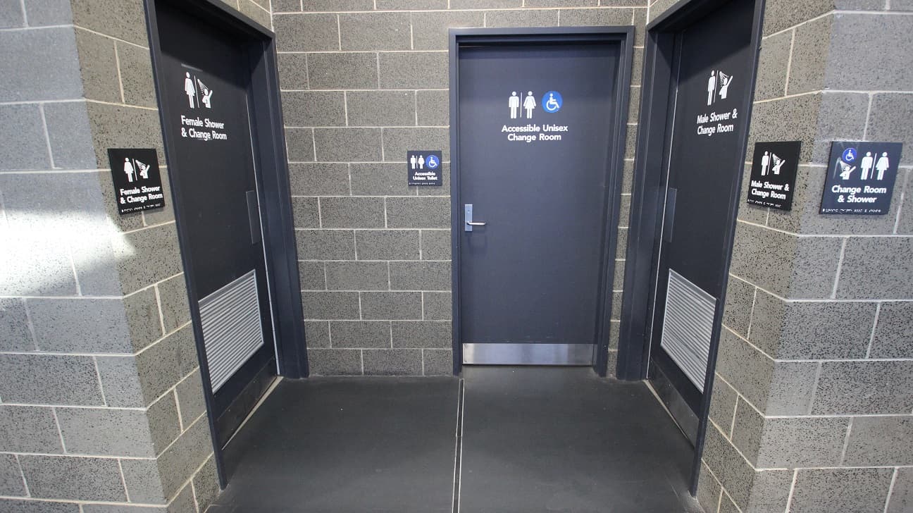 Accessible Unisex Change Room in the middle. It is different from the other changing room due to the blue and white accessible sign beside the male and female icons. On the opposite sides of the accessible unisex change room is the female shower and change room on the left and the male shower and change room on the right. Similar to the corridor walls of the previous image, this also has grey brick walls.