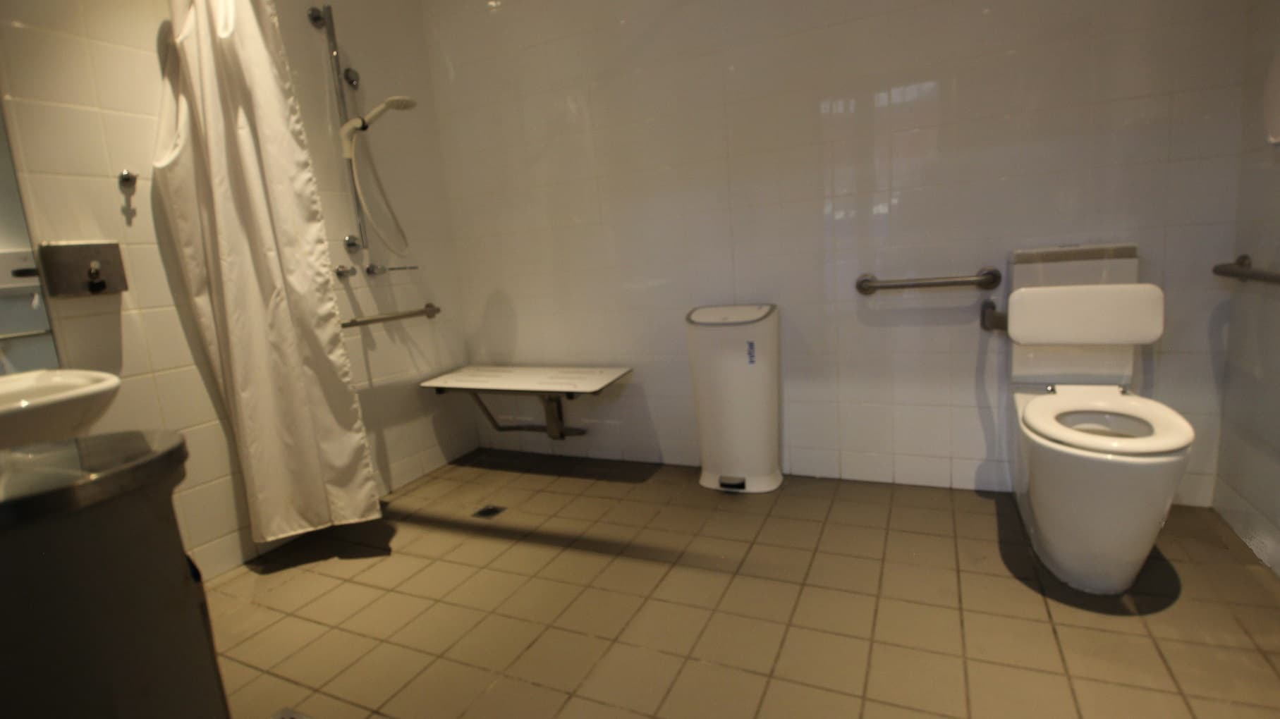 Inside view of one of the Change Rooms at Michael Clarke Recreation Centre. There is a white, hand-held shower, a white seating platform mounted on the wall, and a white privacy curtain during the shower. A wash basin, mirror, and rubbish bin are also available.