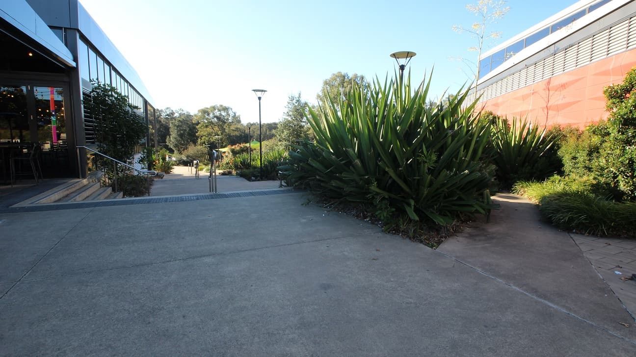 Access to the Tennis courts at Michael Clarke Recreation Centre is via an external pathway. Navigating away from the reception to the right is a sequence of steps and ramps to move down the hill beside the facility. Some noticeable parts of this external path are a lamp post and tree with a white L-shape cemented section.