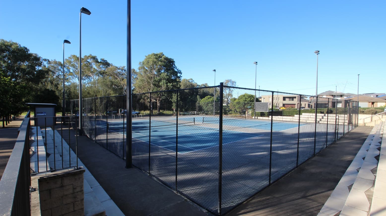 Overlooking the Tennis Courts at the Michael Clarke Recreation Centre from the upper deck of the concrete seating area. Securing the perimeter of the court is a black fence with square holes. Lamp posts are also placed within the black perimeter fence to provide light during the night. Further to the back are tall trees that give shade to the ecstatic audience.