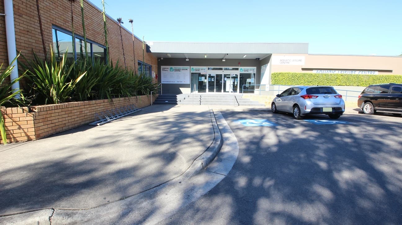 Concrete pathway leading to the four-step grey stairs with three metal handrails to the entrance of the Michael Wendon Aquatic Leisure Centre. Accessible car parking spaces can be found close to and directly opposite the entrance doors. These doors are lined with white edges. In this image, the plants with long leaves in the garden to the left covered the glass windows and had a long stem at the center leveling the topmost part of the wall.