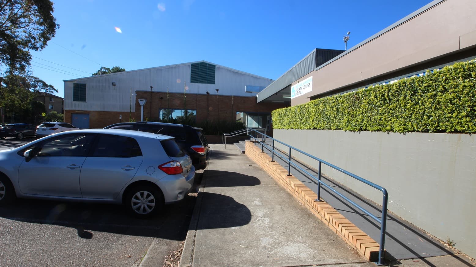 Image shows a concrete pathway before the grey stairs and the accessible ramped access at Michael Wendon Aquatic Leisure Centre. There are tall trees with significant foliage that provide shade to the cars at the parking spaces close to the entrance. From this side, a part of the venue with a white-painted wall and lines can be seen behind the brick wall. It has two windows for air ventilation.
