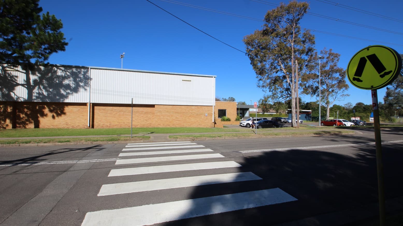 Pedestrian lane leading to the left side of the Michael Wendon Aquatic Leisure Centre. It can be identified by the neon green pedestrian crossing sign containing a walking leg icon and shoes. The lane can be identified by the white Zebra lanes. Cars of various makes and colors are parked in the area in front of the venue. The parking spots closest to the entrance are shaded by tall trees with ample foliage.