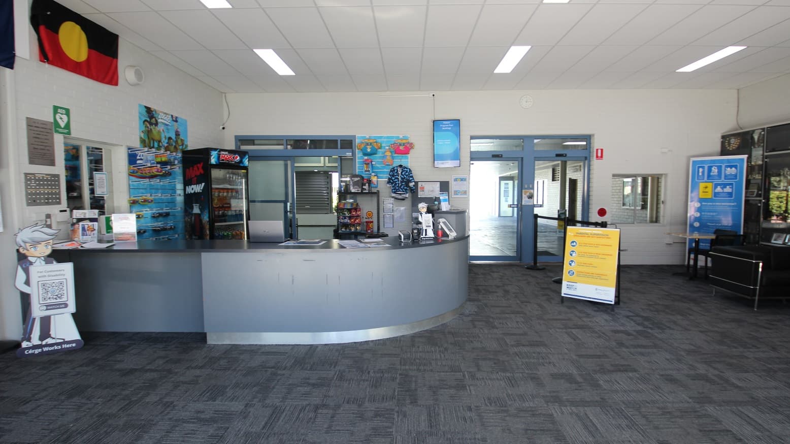 The automatic glass sliding doors at Michael Wendon Aquatic Leisure Centre can be easily distinguished by their white painted edges and grey walls. White square LED light panels are placed on the ceiling. The name of the venue can be found on both upper sides of the entrance doors as well as small and large marketing posters scattered on the glass doors and on the smooth concrete grey wall.