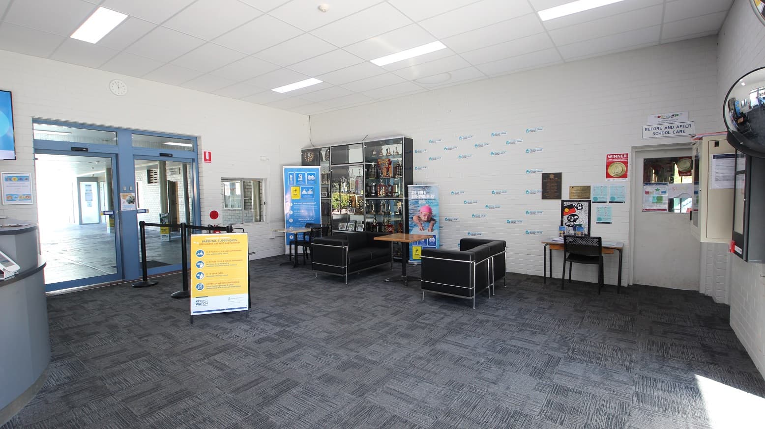 Opposite the curvy reception desk at the Michael Wendon Aquatic Leisure Centre is a seating area that contains black sofa chairs with a brown table in the middle. A grey cabinet storing lots of trophies is covered with glass. Several tickers can also be seen on the white-painted brick wall. On the right is a white door for “before and after school care” while on the left is a black stanchion with black ropes leading to a glass sliding double door with blue painted edges.