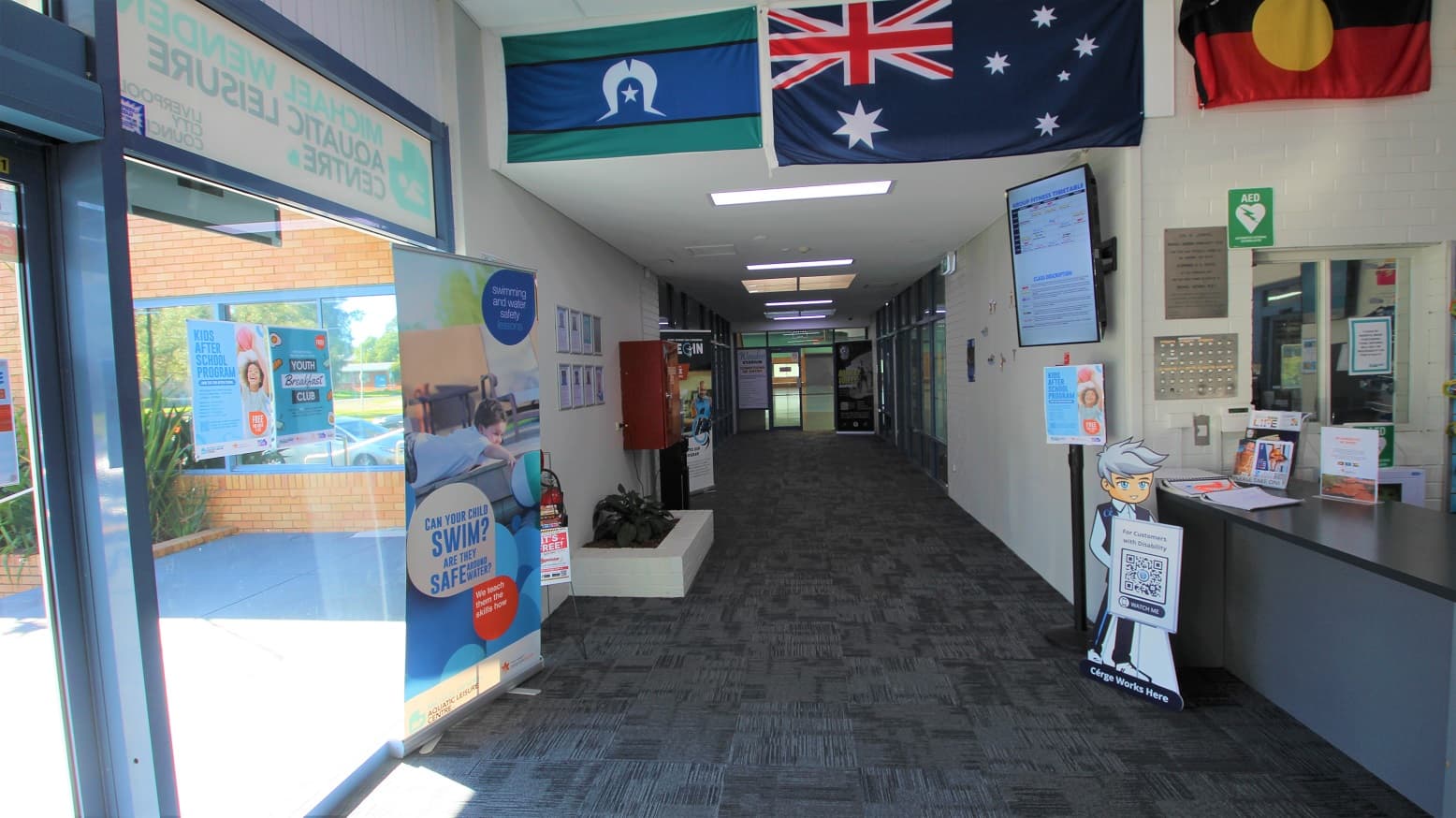 Hallway leading to the Stadium and Gymnasium areas at the Michael Wendon Aquatic Leisure Centre. In front of the reception desk is the brand icon of Cérge with a QR code to help customers with disabilities. The space before the ceiling is occupied by the Australian, Torres Strait, and Indigenous Australian flags. The television mounted on the right wall is in a vertical manner. On the left of this hallway, you can find a fire extinguisher and a small white plant box.