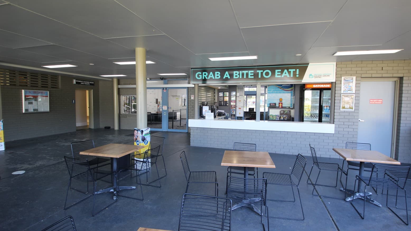Café inside the Michael Wendon Aquatic Leisure Centre. Noticeable in the Café is the signboard which contains a neon blue, “GRAB A BITE TO EAT!”. The brick walls and floors are grey. On the left side is the entry point to the women’s change room and toilet beside a bulletin board. The seating area in the Café is composed of black metal frame chairs and wooden tables with grey support.