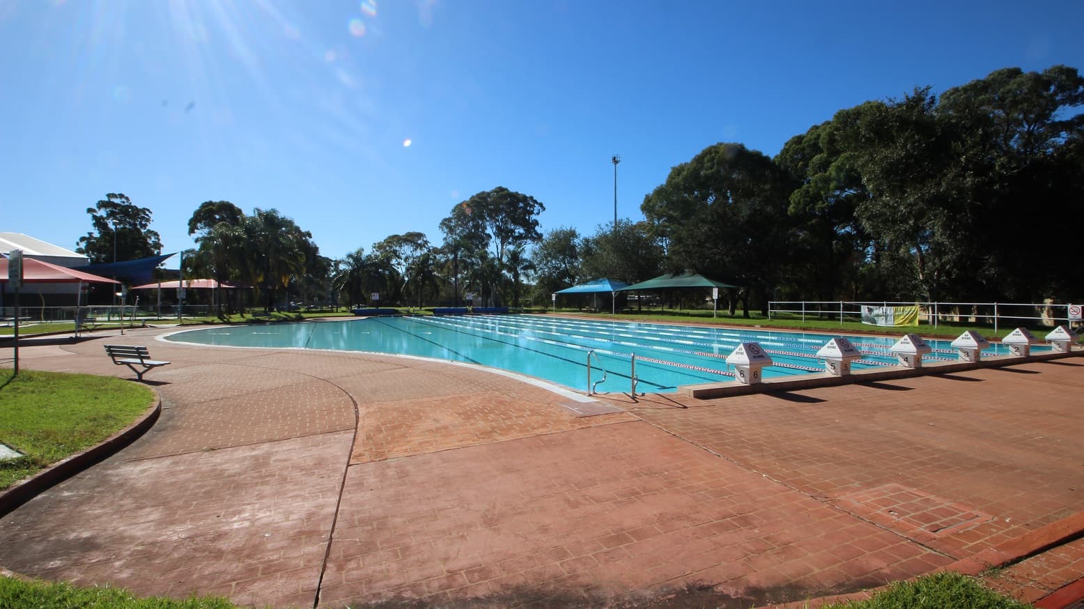 A closer view of the 50 meters outdoor pool located in the park-like outdoor area at Michael Wendon Aquatic Leisure Centre. The lanes are separated by white, blue, and red floating ropes. Surrounding the pool is a brick pathway to move around the area. On the left is a pool ladder. At the top of the slightly elevated section of the pool starting point is a mini-house-looking shape with a no-diving sign and a number etched on the bottom that increments from right to left.