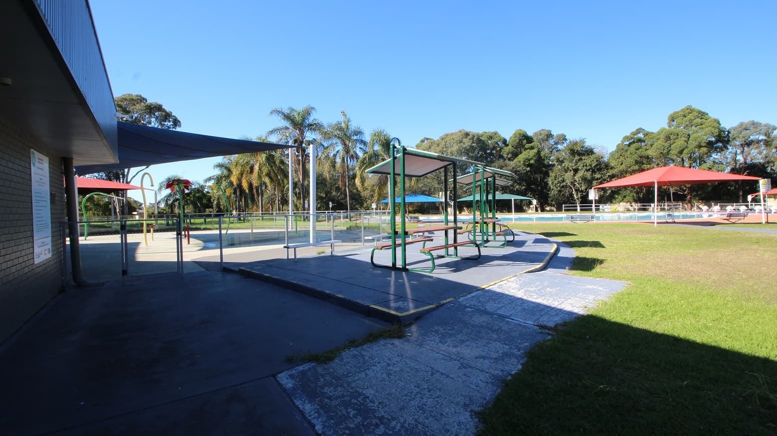 A closer look at the outdoor shelters near the 50 meters outdoor pool. It has a wooden table and seating, white roofing, and a green-painted metal frame. Behind the outdoor shelters is a hip-high glass fence with metal handrails enclosing a space that has yellow and green curvy tubes and bollards.