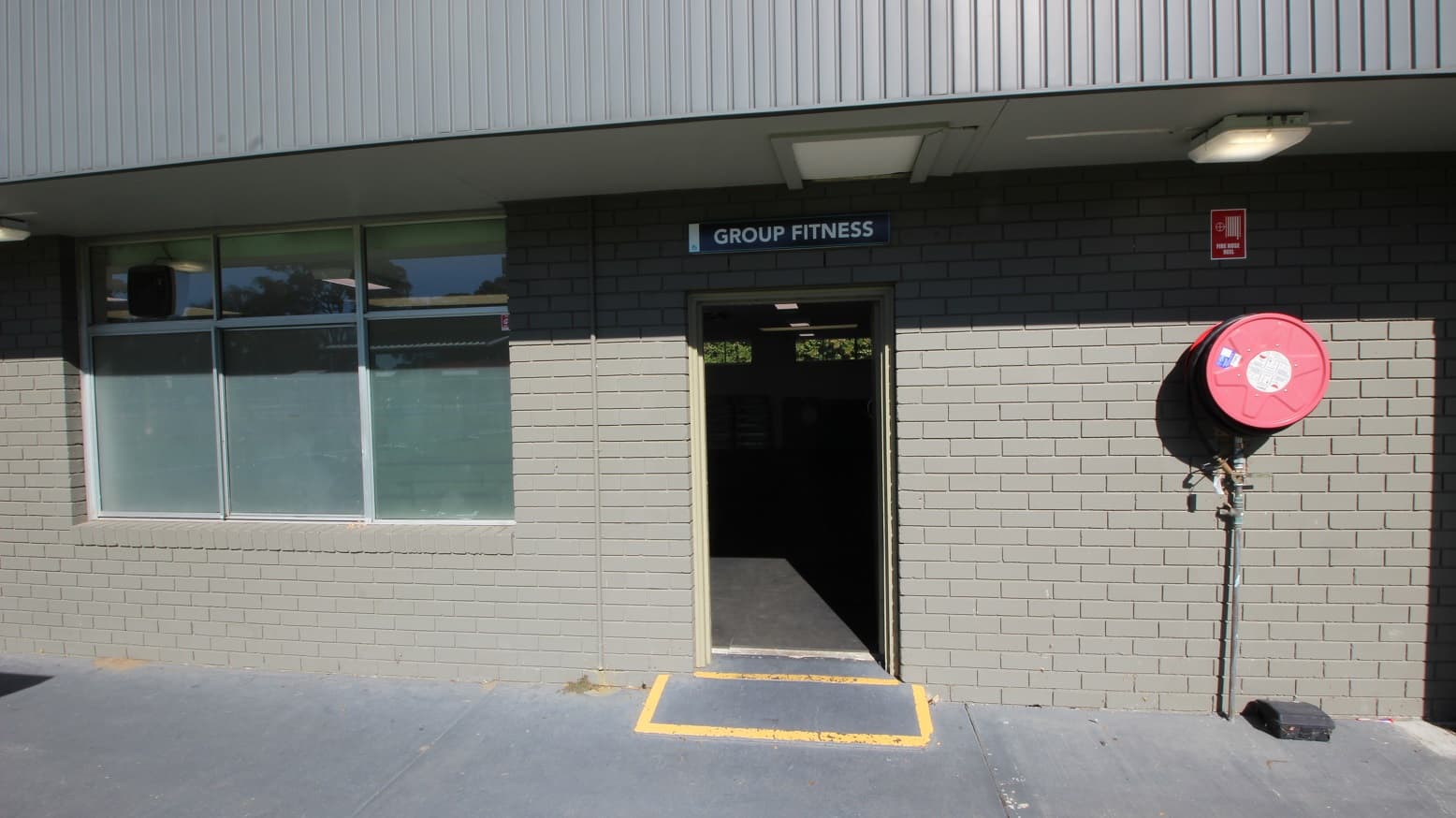 The entrance door to the Group Fitness room at Michael Wendon Aquatic Leisure Centre can be identified by a yellow rectangular marking drawn on the floor. On the right wall is a red fire hose mounted on the grey-painted brick wall. There is a glass window on the left.