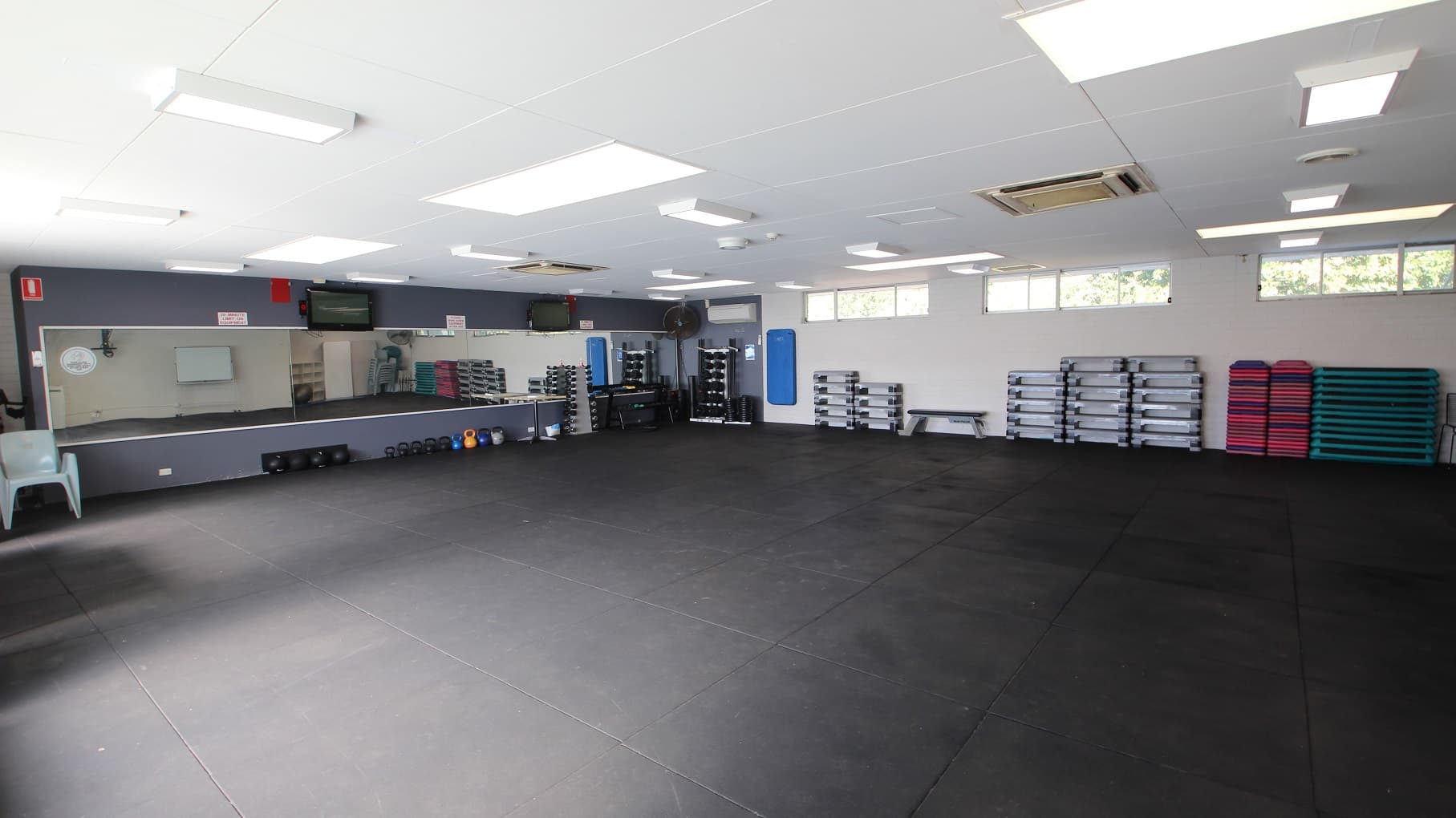 The Group Fitness room has a large open space with a grey rubber mat. The front wall with a medium size mirror is painted grey while the other walls are painted white. On the floor underneath the mirror are body bars in different colors while on the left side is a large white chair. On the upper part of the white-painted wall are small, sliding glass windows and a set of grey platforms stacked on top of each other.