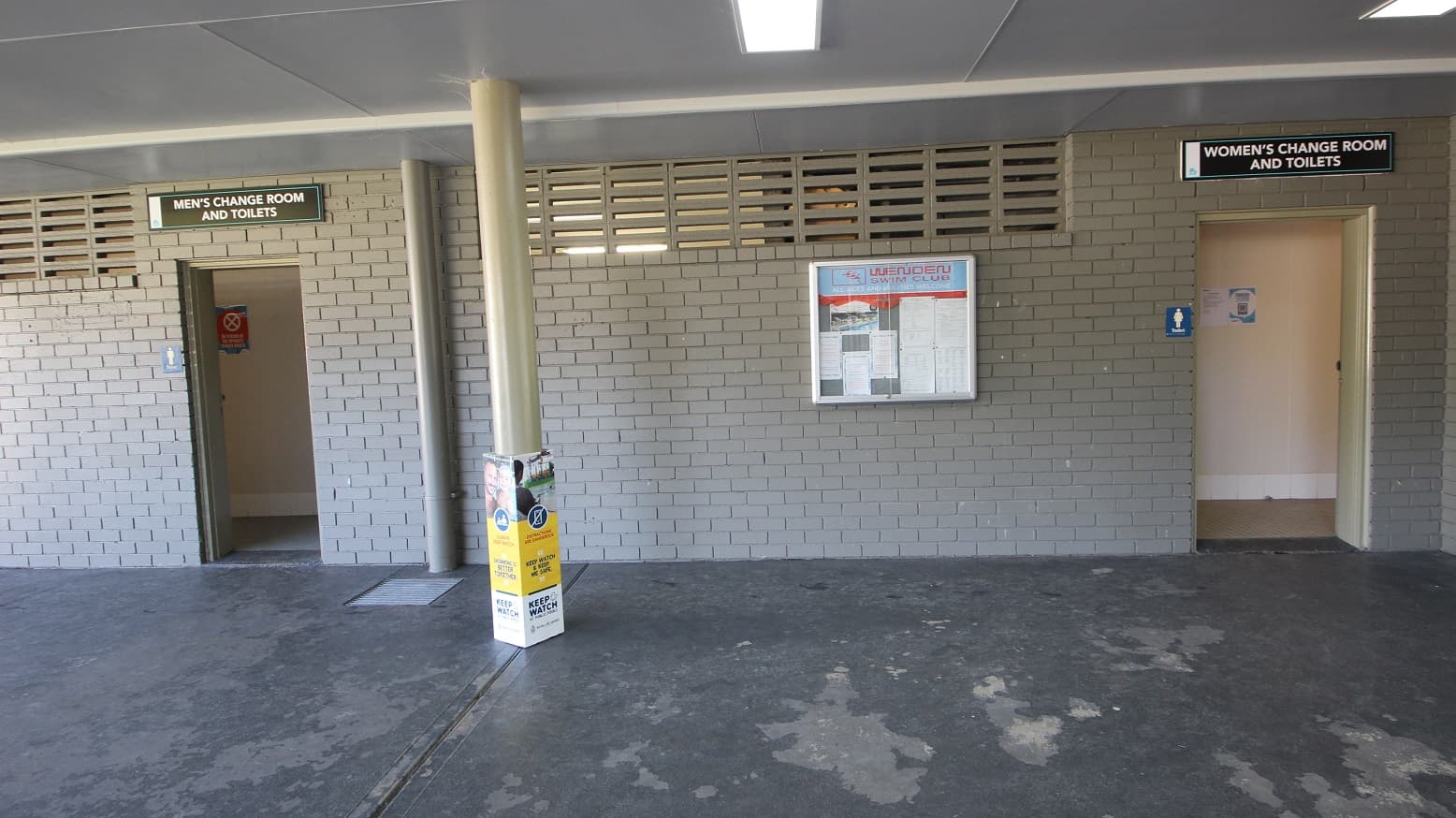 The image shows the men’s change room and toilets on the left side while the women’s change room and toilets are on the right. The brick wall is painted grey and on some parts, on the upper side of the wall, is a column of narrow rectangular holes. Pinned inside the bulletin board located between the wall of the toilet doors are white papers with fine prints.