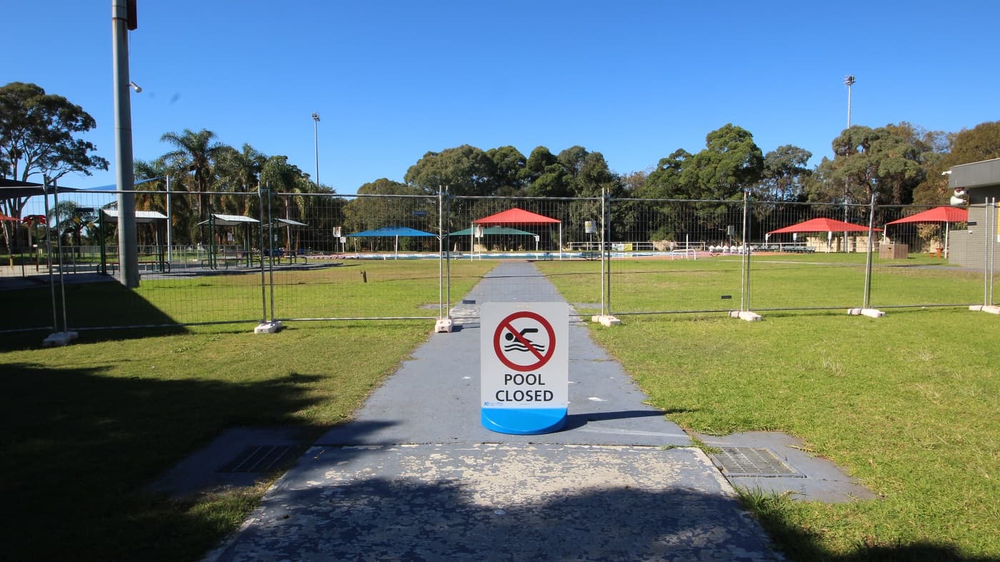 Pool closed sign board placed on a grey, smooth concrete pathway leading to the 50 meters outdoor pool. The area is surrounded by a tall metal fence with small square holes, which gives guests a view of the surrounding area. The large tents, outdoor shelters, palm, and other tree types are visible in this area.