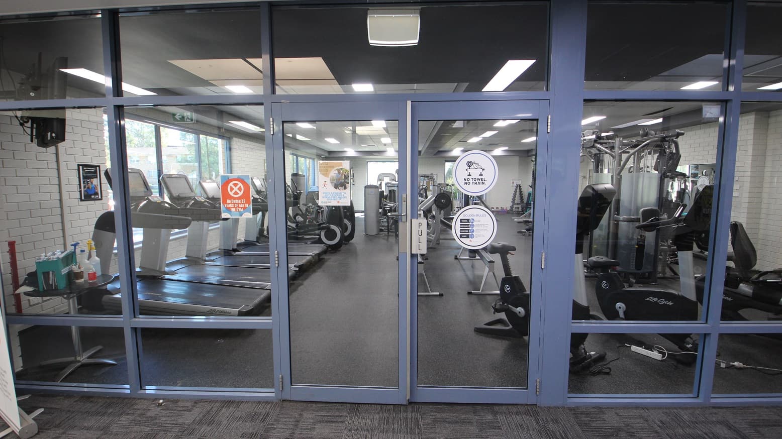 Front of the manually operated glass doors with blue painted edges of the Gymnasium at the Michael Wendon Aquatic Leisure Centre. Opening the door is by pulling it towards yourself. A warning sign on the orange poster on the left prohibits those under 16 years of age from entering this area. Rules and conditions before entering the gymnasium can be found on the right of the entrance door. Seen through the glass doors are cardio and weight training equipment.