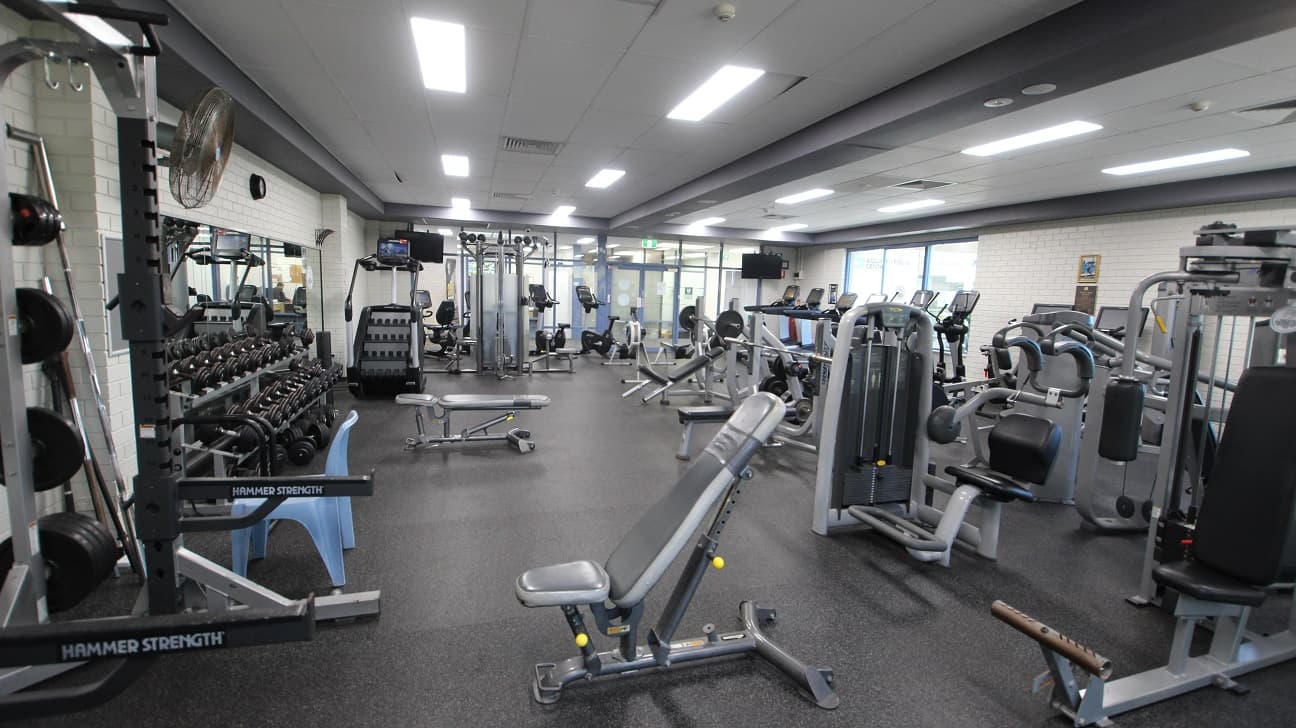 Opposite view inside the gymnasium. This image shows a closer look of the weight training equipment available inside the Gymnasium at Michael Wendon Aquatic Leisure Centre. There is a mirror behind the dumbbell rack and on the upper part is a black analog clock. In front of the wall with a black fan is a large blue plastic chair.