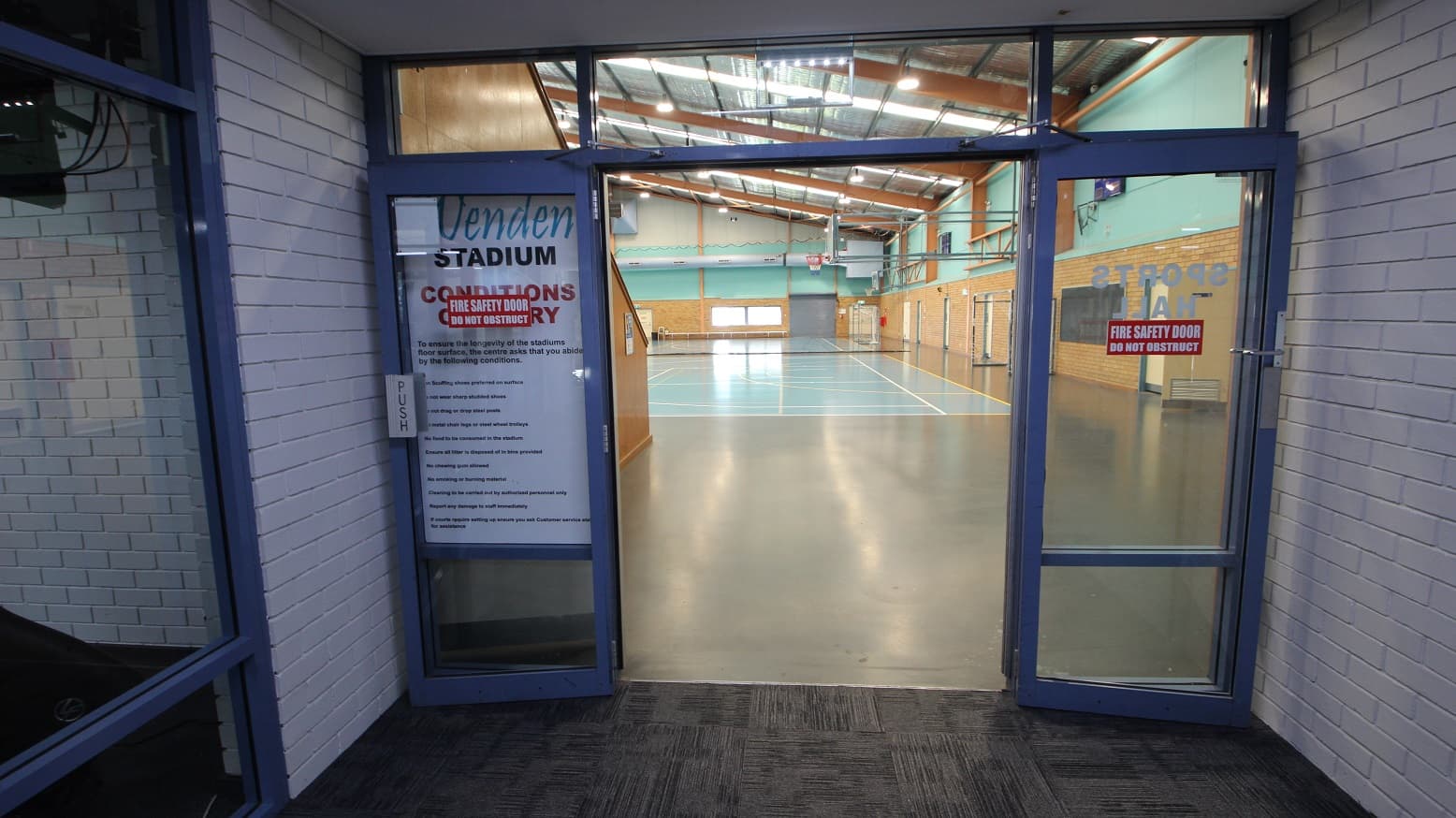 Open doors of the Stadium at the Michael Wendon Aquatic Leisure Centre. The Stadium has a manually operated, glass double door with blue painted edges. The right portion of the Stadium seen through the entry doors shows a sky-blue concrete floor with white & yellow lines. The lower section of the wall is made up of brown-painted bricks, while the upper section is painted sky blue. The Stadium has an insulation-lined roof with skylights and apricot-colored beams.