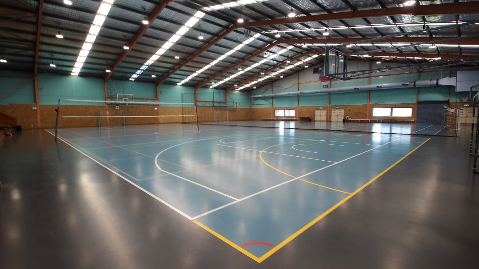 Entire view of the Stadium at the Michael Wendon Aquatic Leisure Centre. The sporting stadium is made of two basketball courts in size and is separated by a black net in the middle. The first court near the entrance doors has a volleyball net while on the second court is a white football net. Scattered near the brick walls and glass windows are long benches. There are dirty-white double doors with horizontal metal handles at one end of this stadium.