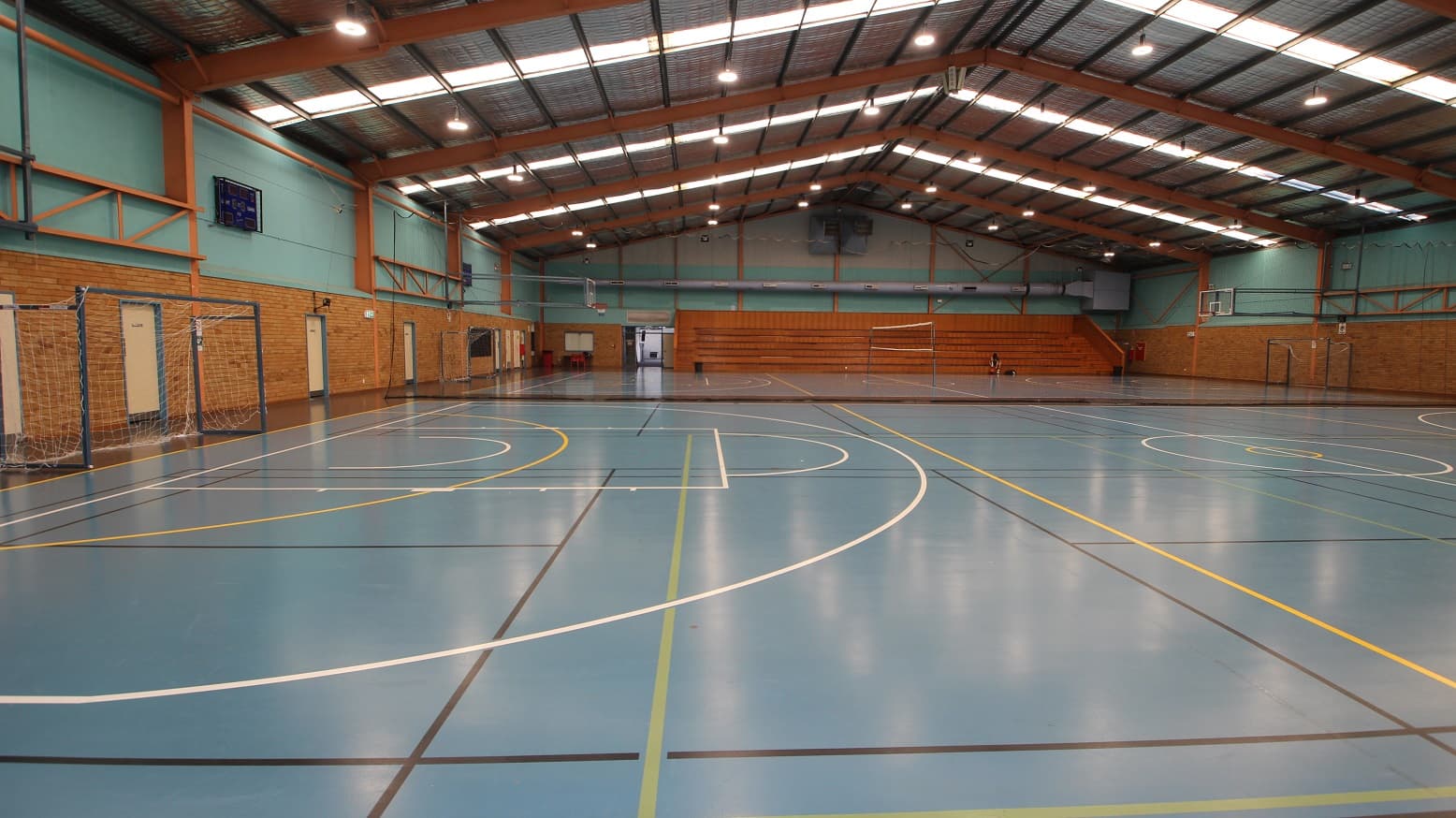 A closer look at the second court of the Stadium at the Michael Wendon Aquatic Leisure Centre. The white football net is wrapped in a grey metal frame. Aside from the white and yellow lines, there is also a black line visible on the floor of this court. There is a six-tiered stadium seating made out of timber situated to the left of the entrance doors.