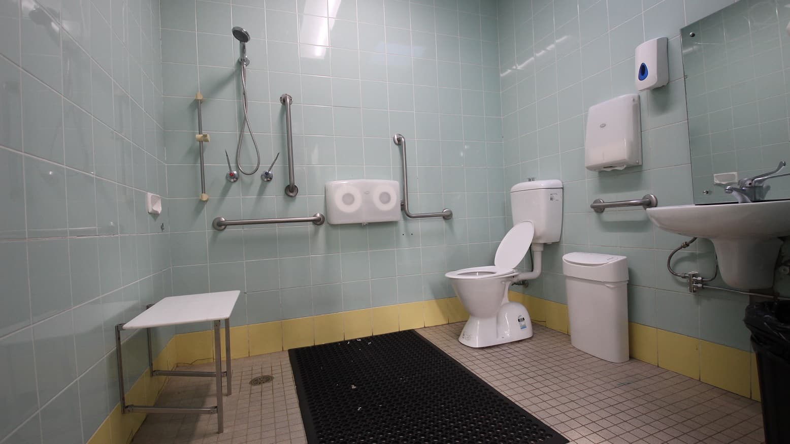 View inside the Accessible bathroom in the Gymnasium at the Michael Wendon Aquatic Leisure Centre. It has a black rubber mat, a wash basin with a mirror, an automatic liquid dispenser mounted on the light blue tiled wall, a white and black rubbish bin, two rolls of tissue papers, a handheld shower, and grab bars. The tiles at the lowermost part of the wall are painted in yellow.