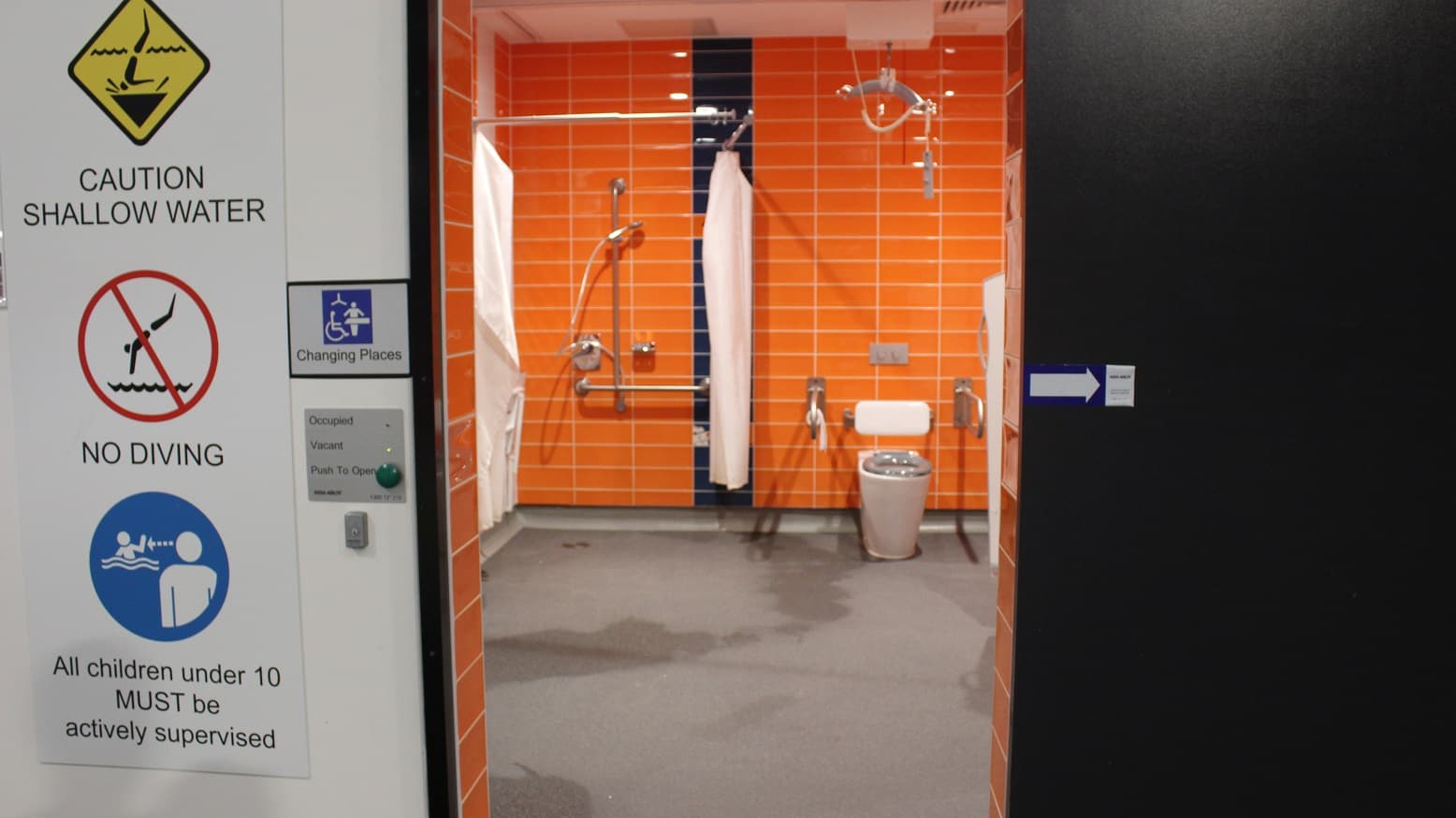 Changing Places at Mill Park Leisure. Left of the entrance door is a No Diving, Caution Shallow Water sign, under 10 children supervision reminder, a push-button for opening, and notification lights for occupied and vacant. Inside the Changing Places is an orange tile wall, a fully accessible toilet, an adult change facility with sensor lighting, grab bars on both sides of the toilet, a sink with lever tap, a shower with HHS, grab bars and shower bench, and a Guldmann ceiling hoist.