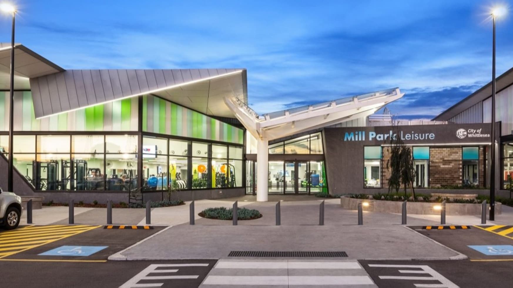 Outside appearance of Mill Park Leisure building with a unique pointed-like roof design facing the same direction. Four parking spaces are available with striped yellow lines and blue bollards separating them.