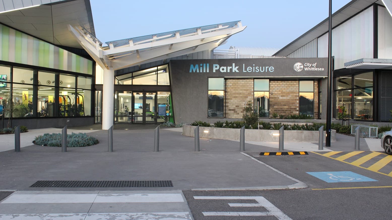 A concrete pathway separates the eleven accessible parking bays leading toward the entrance of Mill Park Leisure. Grey bollards with pointed tops are arranged in front of the parking area. The entrance can be distinguished by the white wing-looking structural roofing with glass on top. Beside the main entrance is a grey building with brick walls and glass windows marked with Mill Park Leisure brand name and the City of Whittlesea and a logo.