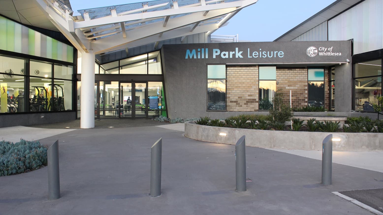 The entrance can be distinguished by the white wing-looking structural roofing with glass on top. Beside the main entrance is a grey building with brick walls and glass windows marked with Mill Park Leisure brand name and the City of Whittlesea and a logo. In front of this is a circular garden with medium-sized ferns.