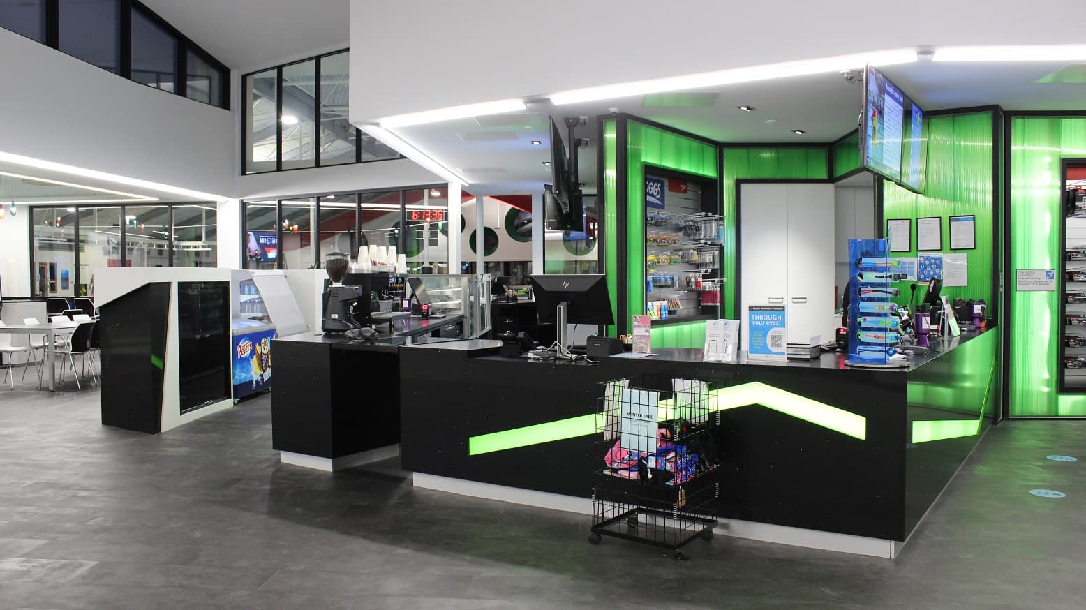 Black reception desks and black Cafe display table area inside Mill Park Leisure. The reception area has neon green walls and neon green lines on its black table and several retail products for sale. Business permits put in a frame are mounted on the neon green walls. Video information can be seen playing on the television sets on top of both the reception and Cafe areas. The front of the Cafe is a seating area with black and white tables and chairs