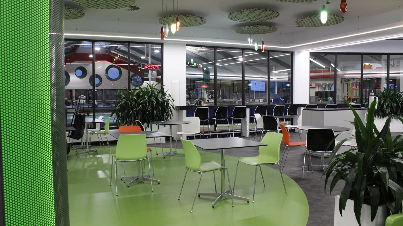 Apart from the black and white tables and chairs in the seating area in front of the Cafe are the green and orange chairs with steel legs. Potted plants are strategically placed around the seating area. Colorful bulbs with tear shapes and circular flat baskets with black and green stripes can be seen on the ceiling. The pool hall is visible through the glass walls.