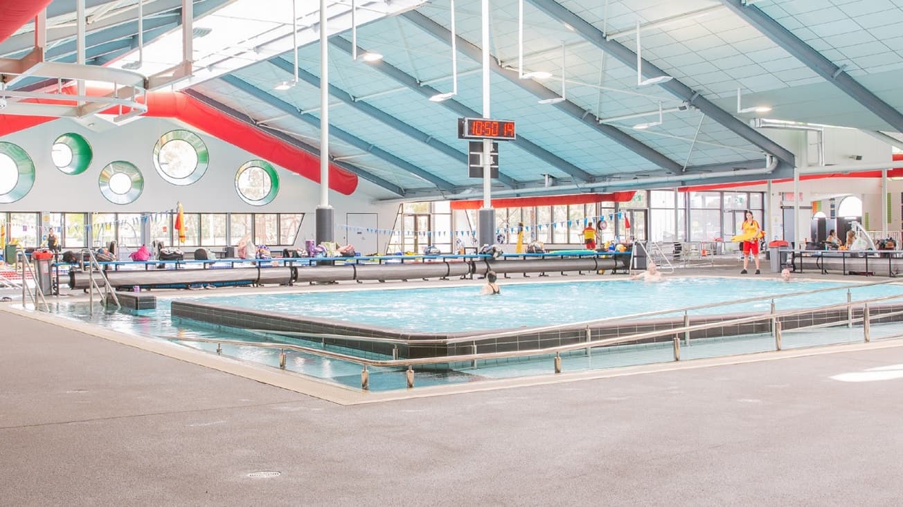 The 23-meter indoor pool at Mill Park Leisure has lifeguards wearing orange and yellow uniforms. In one of the white posts inside the Pool Hall is a large digital clock. Near the pool is a large circular steel tube for sitting and blue tables for bags.