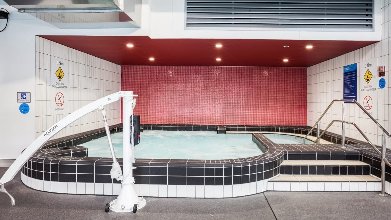 Warm Water Pool/Spa at Mill Park Leisure can be distinguished by the red wall at the back and top and is surrounded by grey tiles. A large white signboard for shallow water and no diving sign is placed on both sides of the walls. On the right side is a metal handrail while on the left side is a white pelican hoist sling to help support and lift persons with mobility issues.