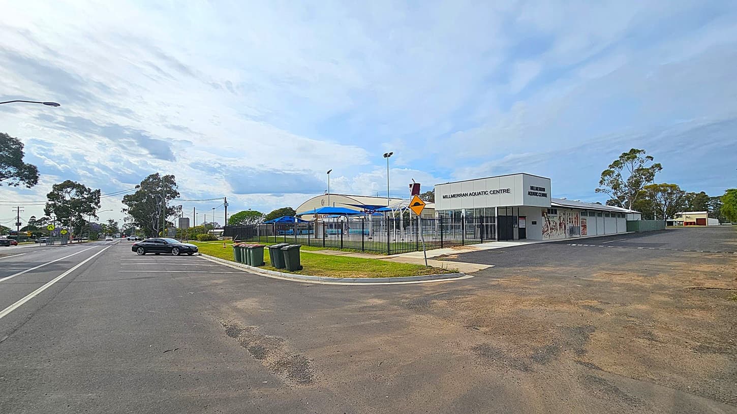 A street view with a parking area on the left, featuring a few parked cars and several rubbish bins. On the right, there's a building labelled "Millmerran Aquatic Centre" with a fenced pool area and blue shade structures. The background includes trees and a few more buildings. The ground is a mix of asphalt and grass, and the sky is overcast.
