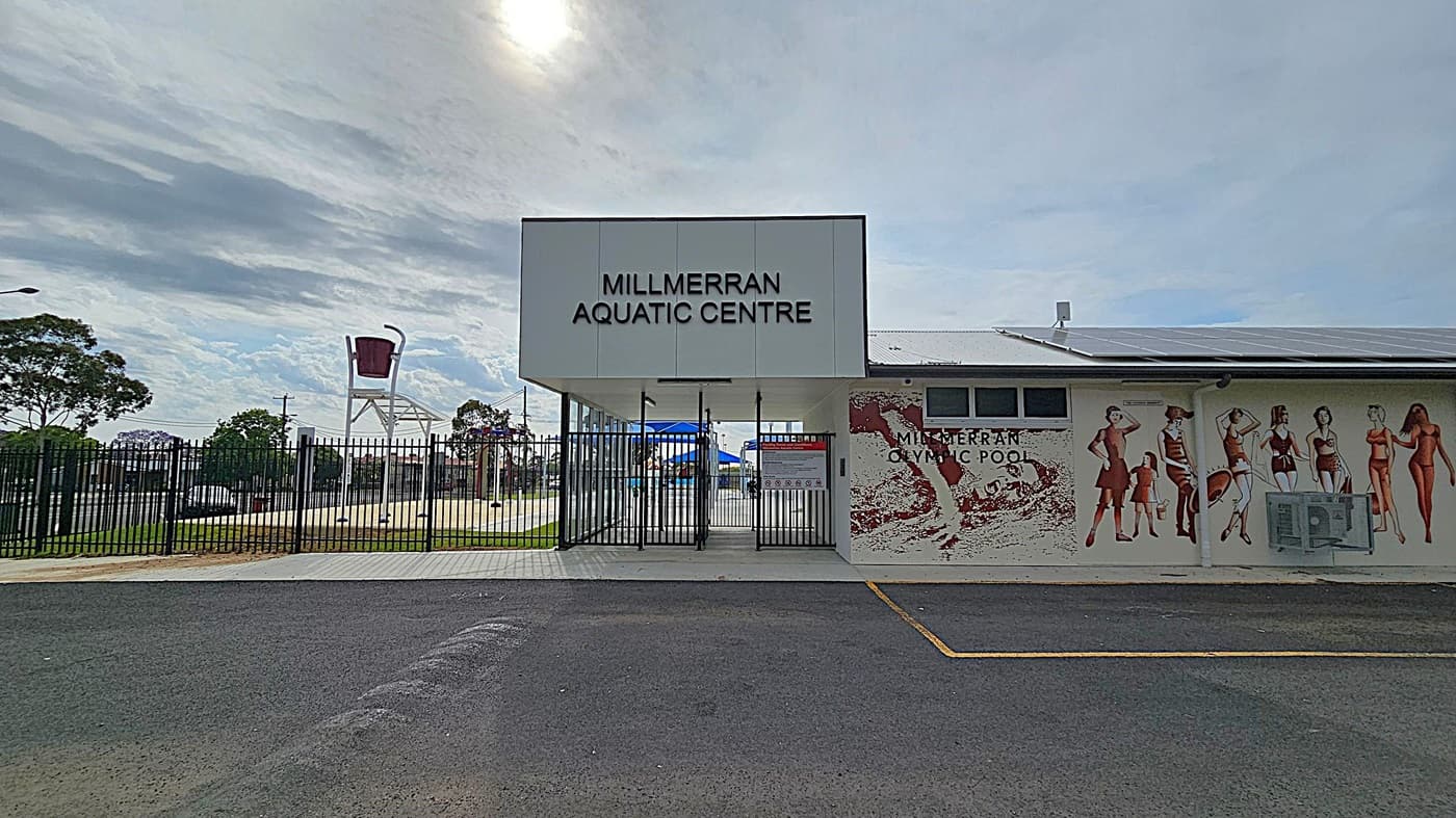 Entrance to the Millmerran Aquatic Centre with a large sign above the gate. To the right, a mural on the building wall depicts people in swimwear. The ground is paved with asphalt, and a black metal fence surrounds the area. In the background, a large bucket feature is visible, likely part of a water play area. Trees and a street are visible beyond the fence.