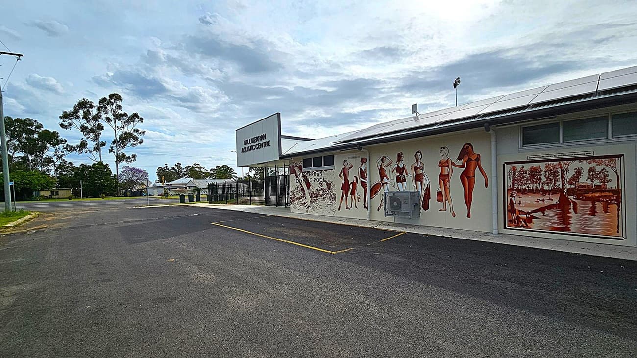A building with a sign reading "Mulgildie Aquatic Centre" on the left side. The wall features a mural of people in vintage swimwear and a painting of a riverside scene. The foreground shows an empty asphalt parking area with yellow lines. In the background, there are trees and a few houses. The sky is overcast.