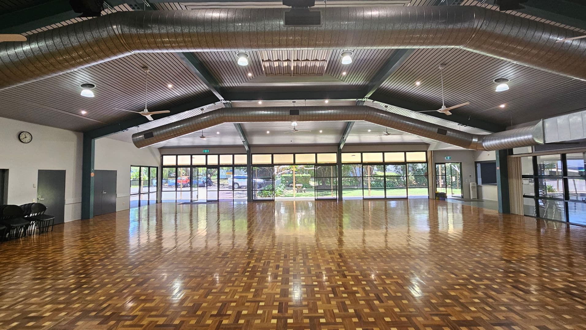 A spacious hall with a polished parquet floor and a high ceiling featuring exposed metal beams and large air ducts. On the left, there are stacked metal chairs against a wall. The back wall has a clock and several doors, with a small table nearby. Large windows on the right allow natural light to enter, showing a glimpse of greenery outside. The ceiling has multiple lights and fans.