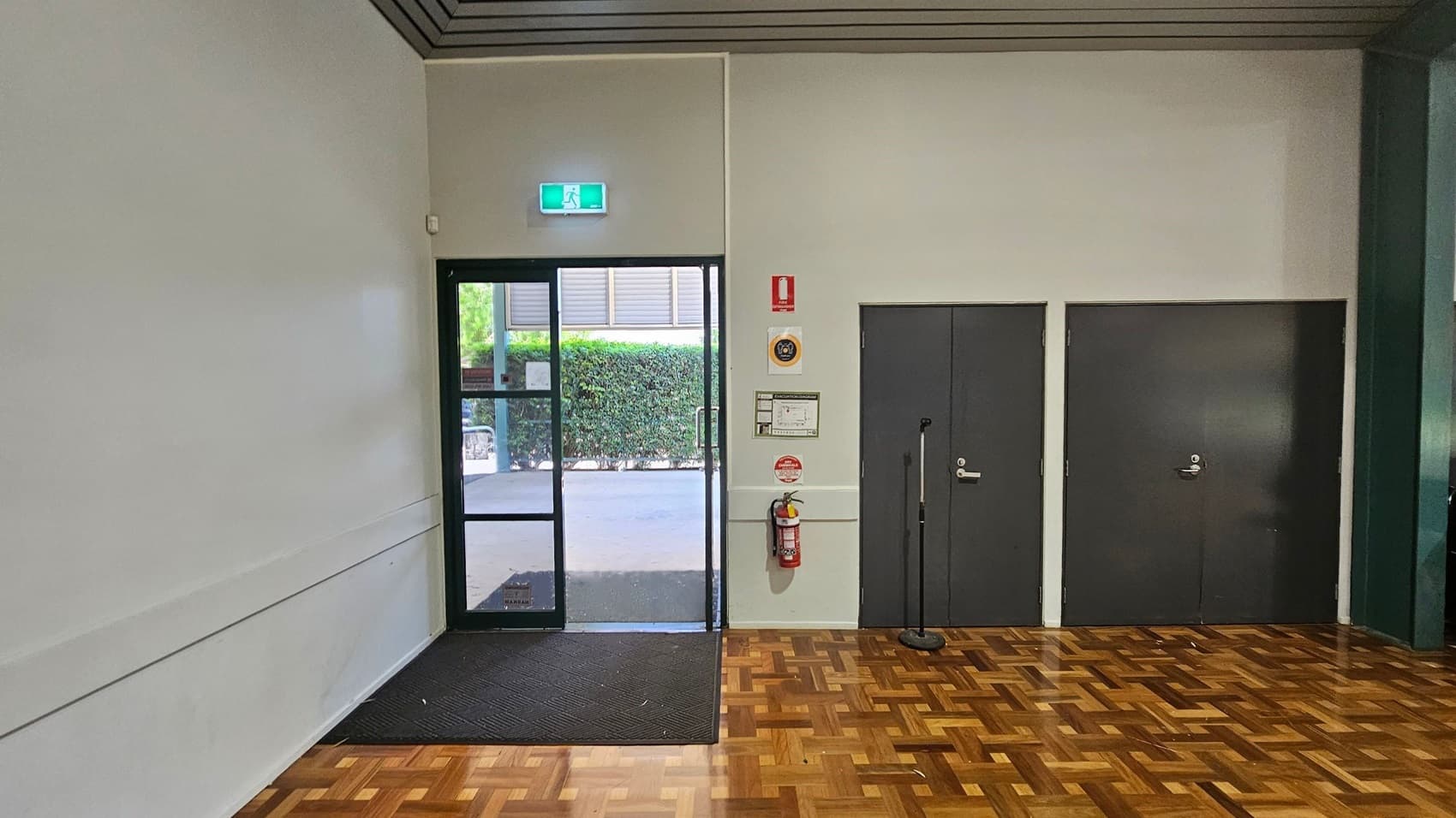 A room with a parquet wooden floor and a light green ceiling. On the left, there's a glass door with an illuminated exit sign above it, leading outside where greenery is visible. To the right of the door, there are two dark grey doors. A fire extinguisher is mounted on the wall between the glass door and the grey doors, along with some safety signs. A black mat is placed in front of the glass door.