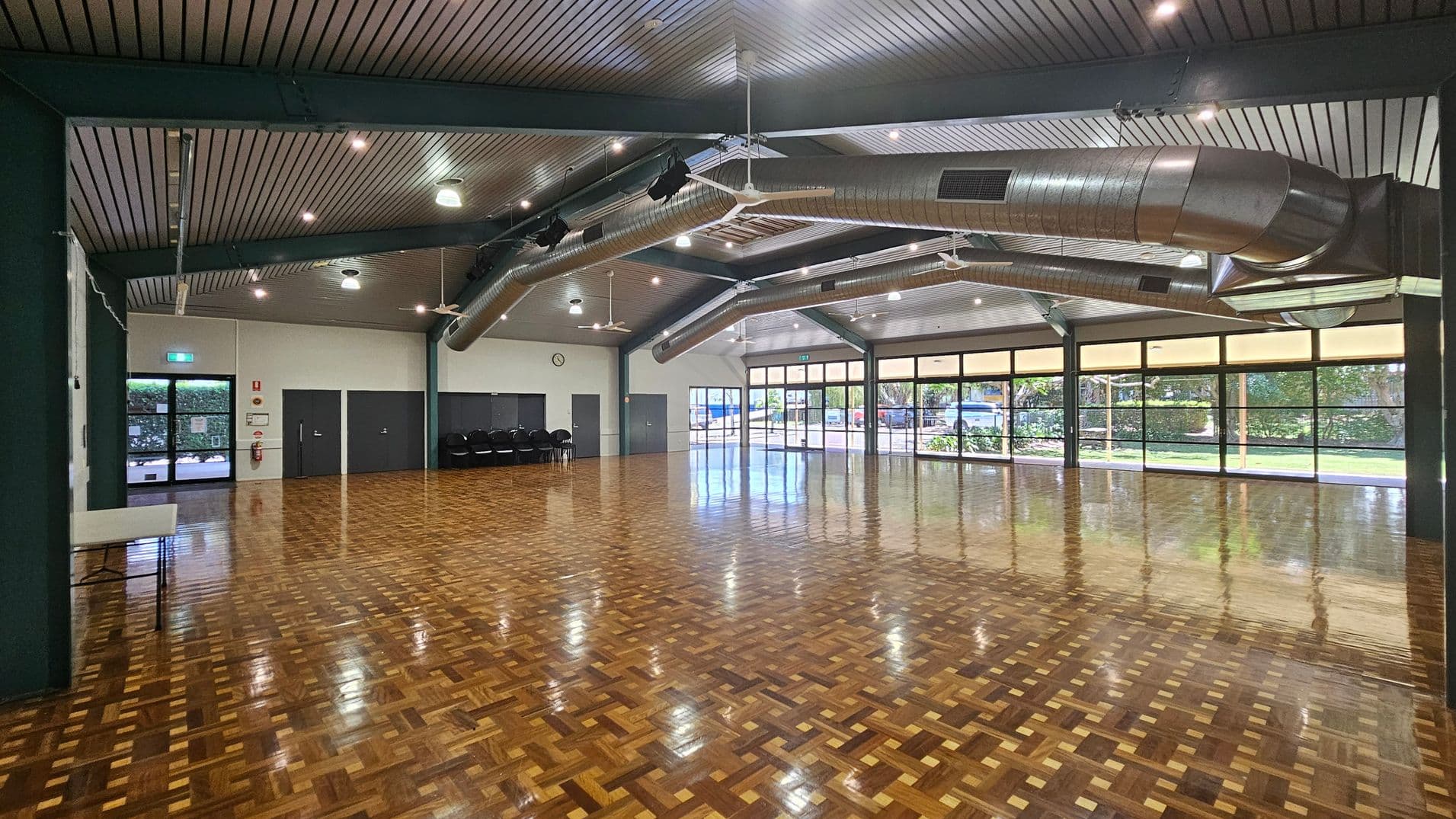 A spacious hall with a polished parquet floor and a high, slatted ceiling featuring exposed ductwork and ceiling fans. On the left, there are dark double doors and a stack of chairs against the wall. The right side has large windows with views of greenery outside. The ceiling has multiple lights, and the walls are light-coloured. A clock is mounted on the wall near the doors.