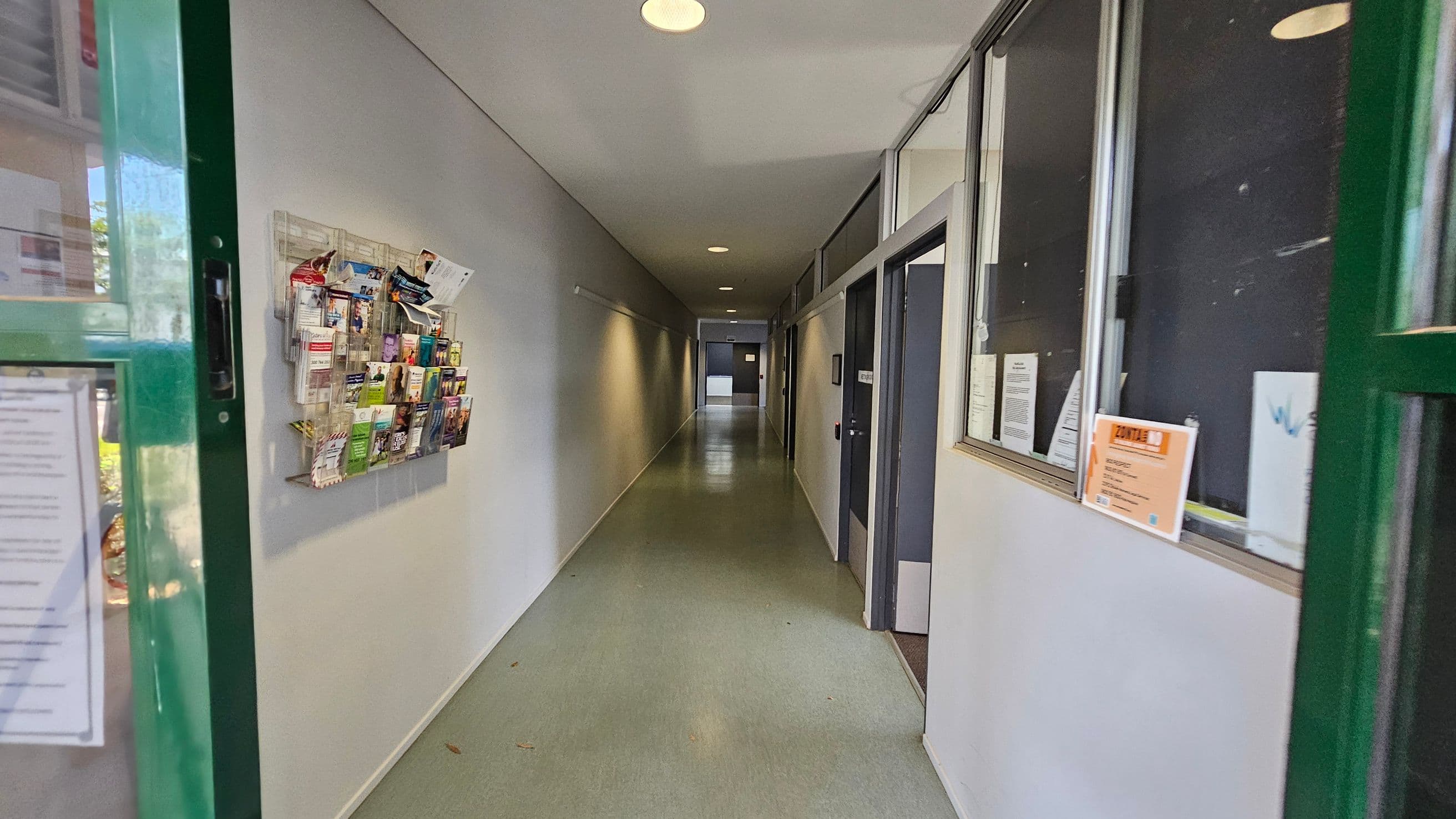 A hallway with a green door partially open on the left. A wall-mounted rack filled with brochures is on the left wall. The floor is light green, and the ceiling has recessed lights. On the right, there are windows with notices and a door. The corridor extends into the distance, with more doors and a light at the end.